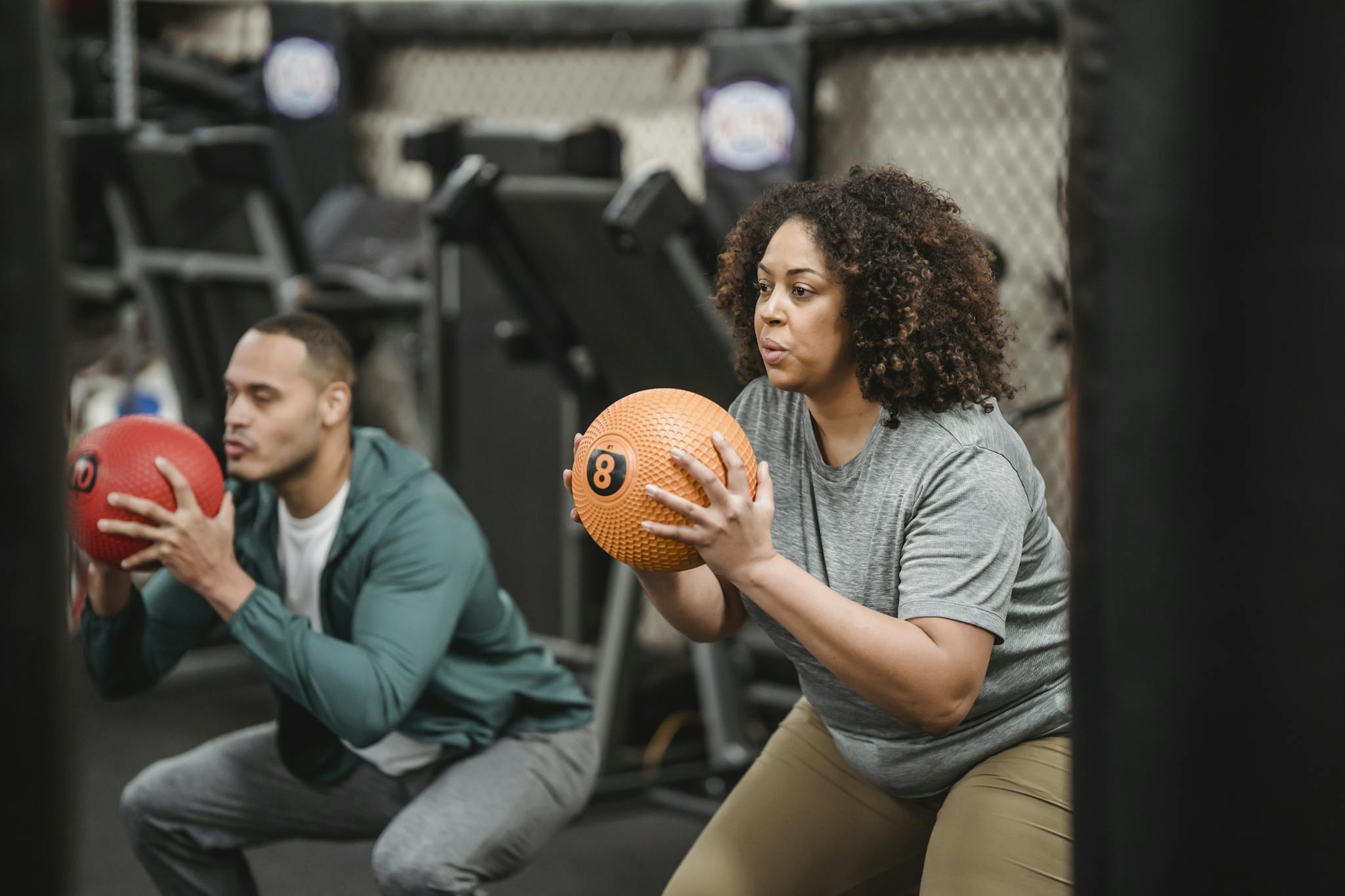 Focused young plus size black female in sportswear doing squats with medicine ball while training in gym with muscular ethnic male trainer